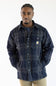 A man wearing a navy blue and gray plaid Carhartt button-up overshirt with a small yellow and white Carhartt logo patch on the chest pocket, paired with khaki pants, against a plain white background.