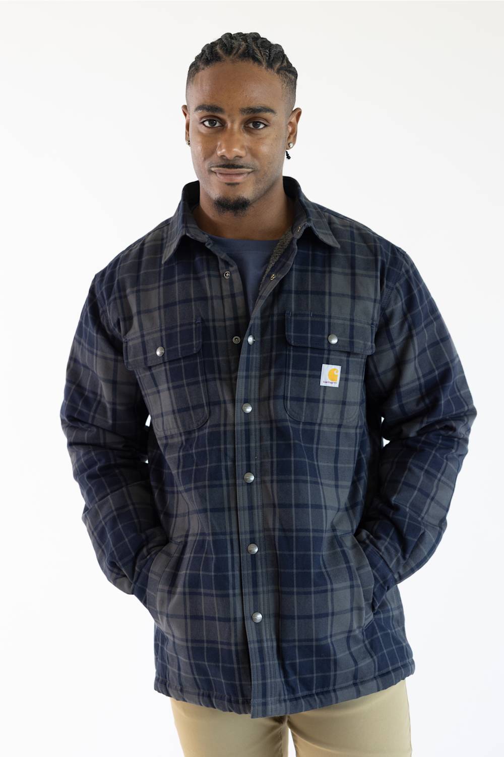 A man wearing a navy blue and gray plaid Carhartt button-up overshirt with a small yellow and white Carhartt logo patch on the chest pocket, paired with khaki pants, against a plain white background.