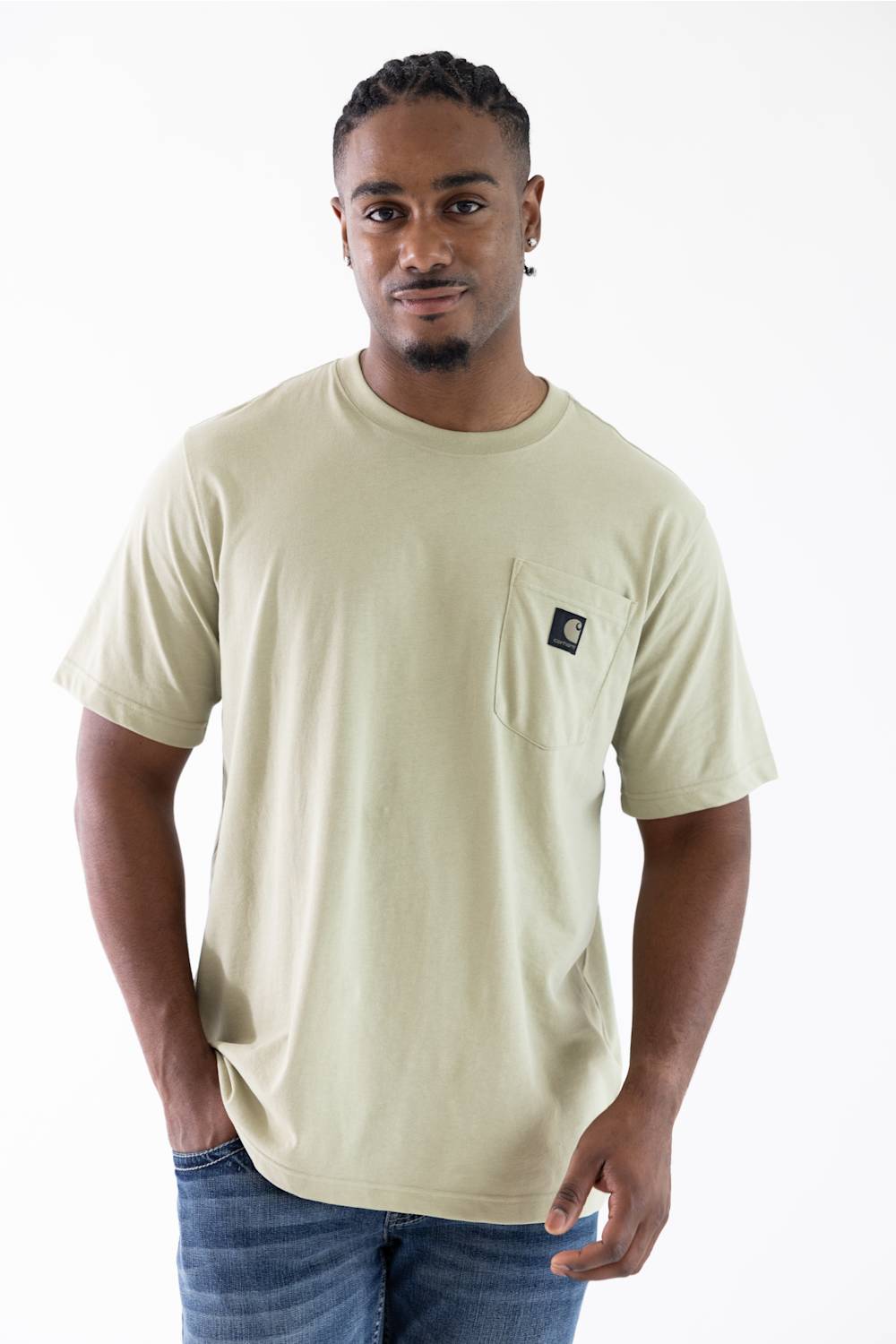 A man wearing a light beige crewneck pocket t-shirt with a small black Carhartt logo patch on the chest pocket, paired with blue jeans, against a plain white background.