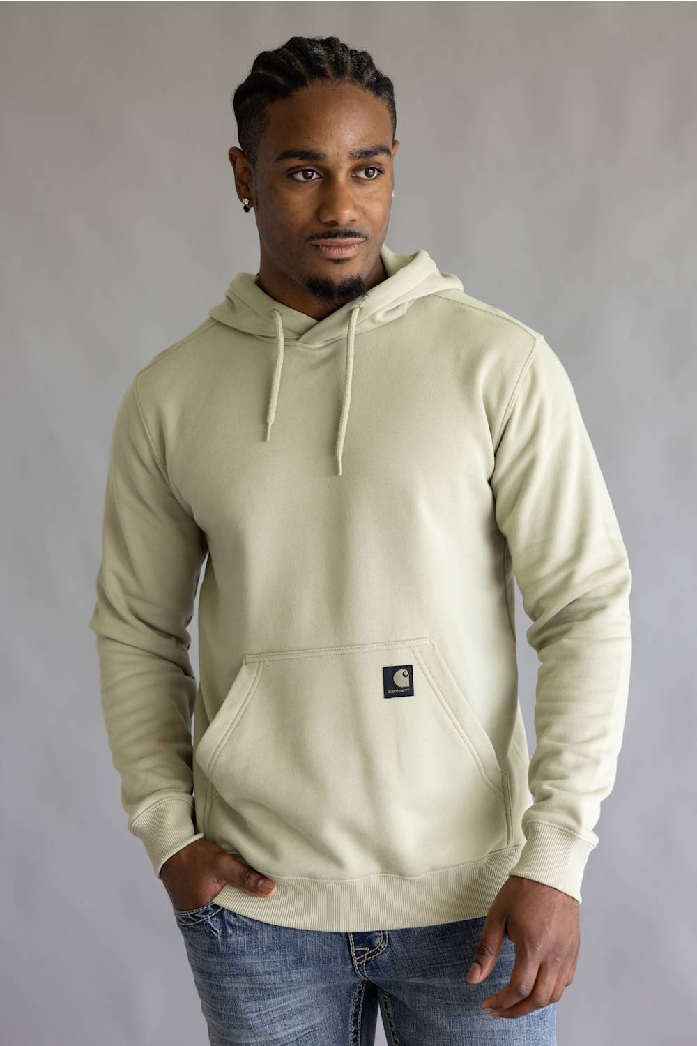 A man wearing a light beige Carhartt pullover hoodie with drawstrings and a front kangaroo pocket, paired with blue jeans, standing against a neutral gray studio background.
