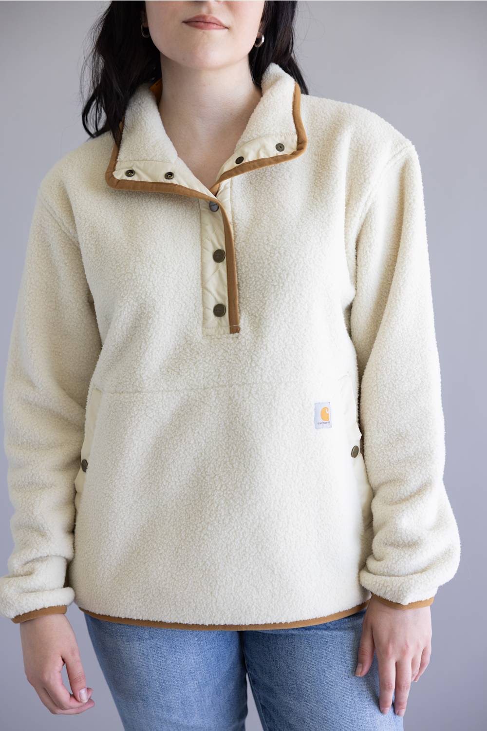 A person wearing a cream sherpa fleece pullover with tan contrast trim and a partial snap placket, paired with blue jeans against a plain light gray studio background.