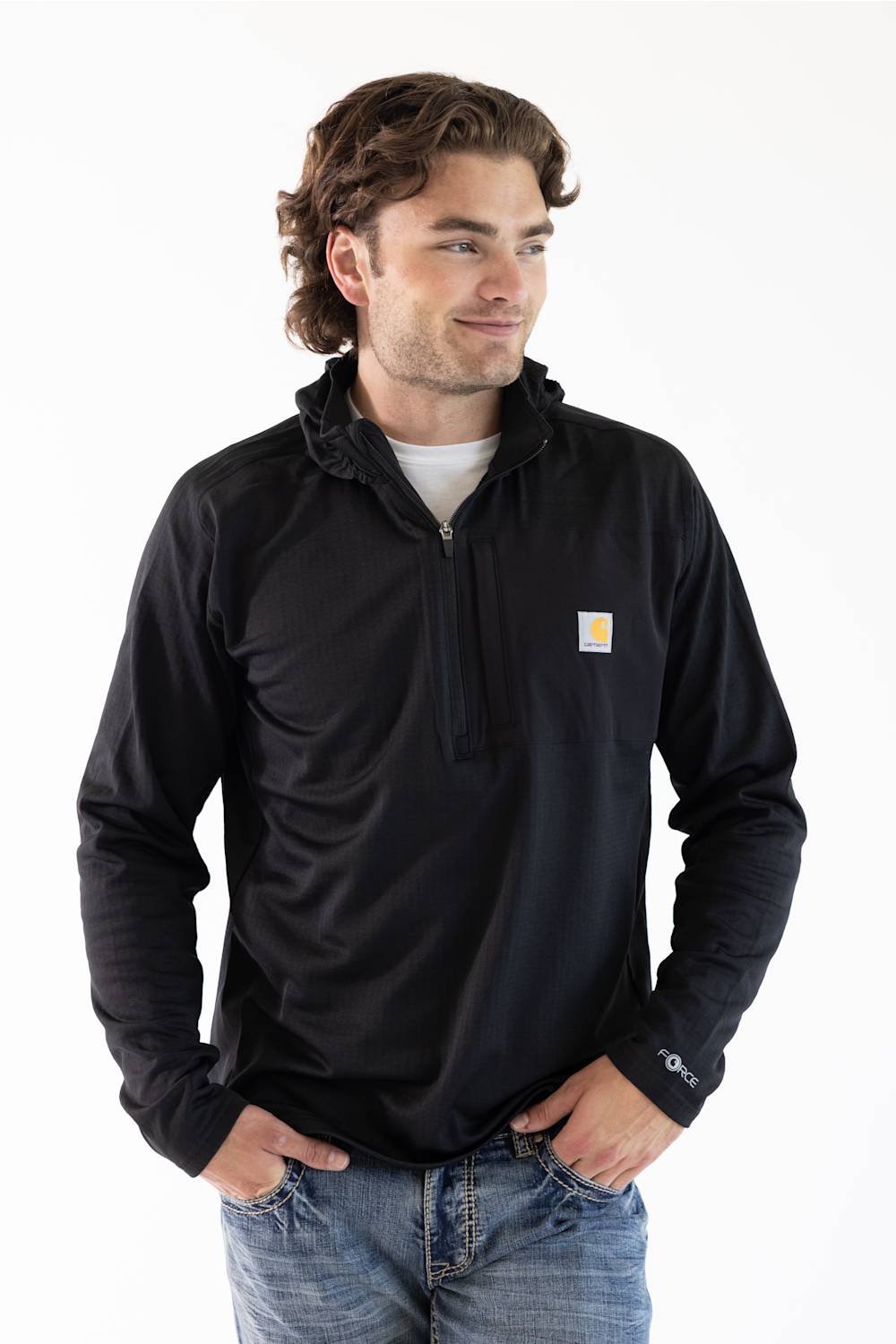 A male model standing against a white background, wearing a black Carhartt Force 1/2 zip hooded sweatshirt over a white t-shirt and blue jeans.