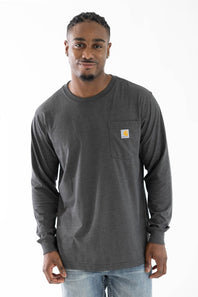 A man wearing a dark charcoal gray long-sleeve pocket t-shirt with a small yellow and brown Carhartt logo on the left chest, paired with light blue jeans, against a plain white background.