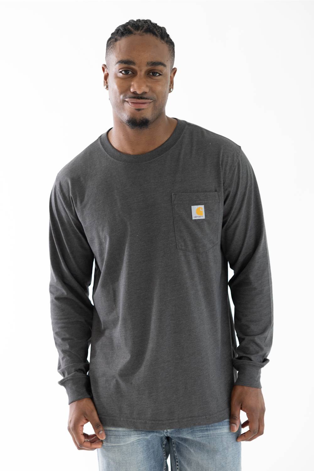 A man wearing a dark charcoal gray long-sleeve pocket t-shirt with a small yellow and brown Carhartt logo on the left chest, paired with light blue jeans, against a plain white background.