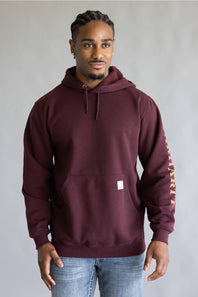 A man wearing a maroon Carhartt pullover hoodie with a small white Carhartt logo patch on the front pocket and vertical gold lettering on the right sleeve, paired with blue jeans, against a neutral gray background.