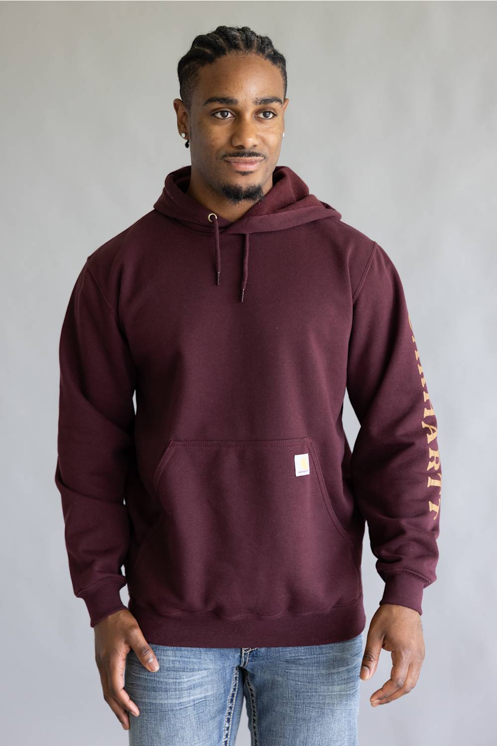 A man wearing a maroon Carhartt pullover hoodie with a small white Carhartt logo patch on the front pocket and vertical gold lettering on the right sleeve, paired with blue jeans, against a neutral gray background.
