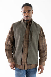 A man wearing a brown and tan plaid button-down shirt under a sleeveless olive green vest, paired with blue jeans, against a plain white background.