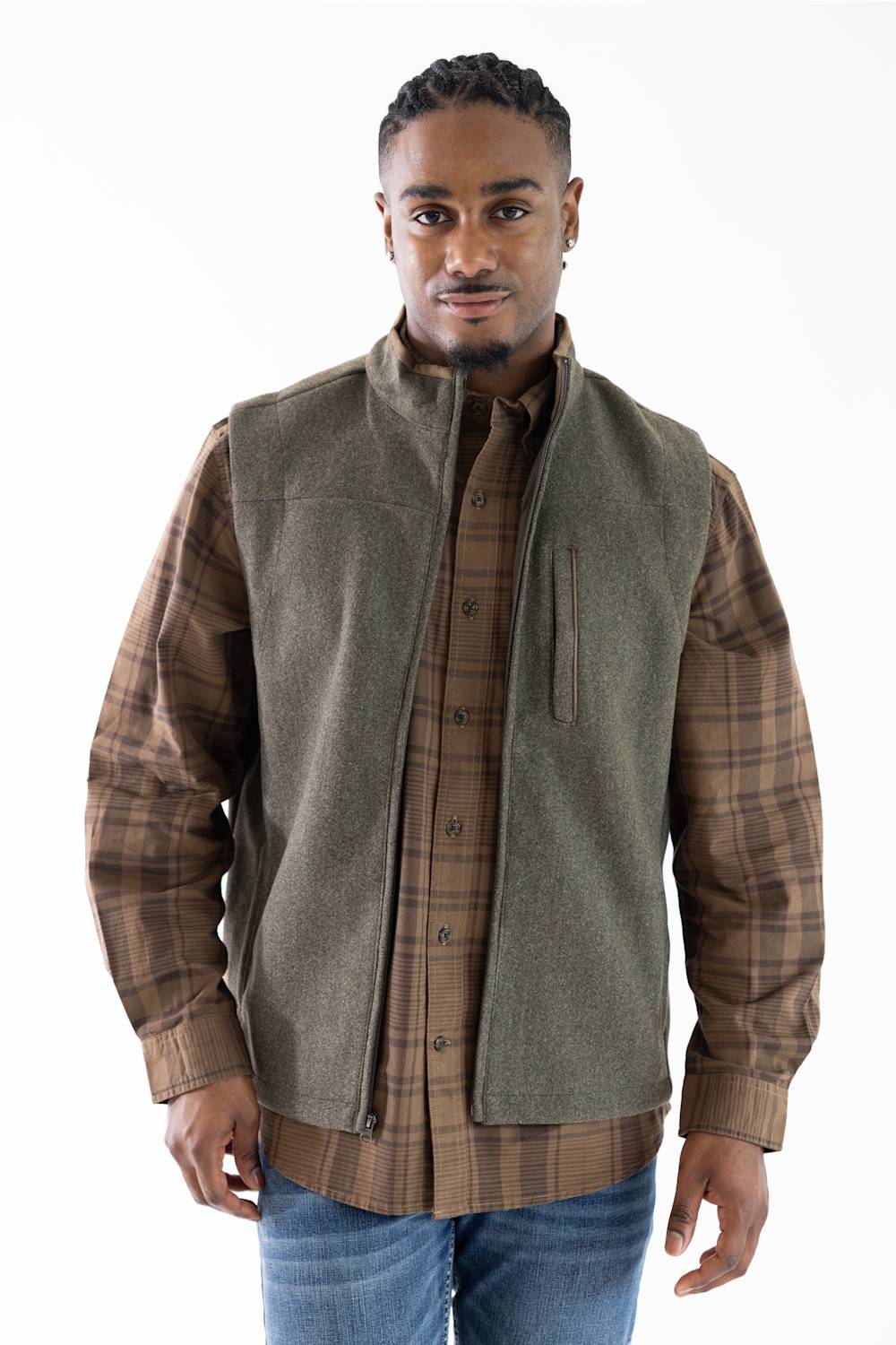 A man wearing a brown and tan plaid button-down shirt under a sleeveless olive green vest, paired with blue jeans, against a plain white background.