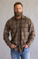 A man with a full beard wearing a brown and beige plaid button-down flannel shirt and blue jeans, standing against a neutral gray studio background.