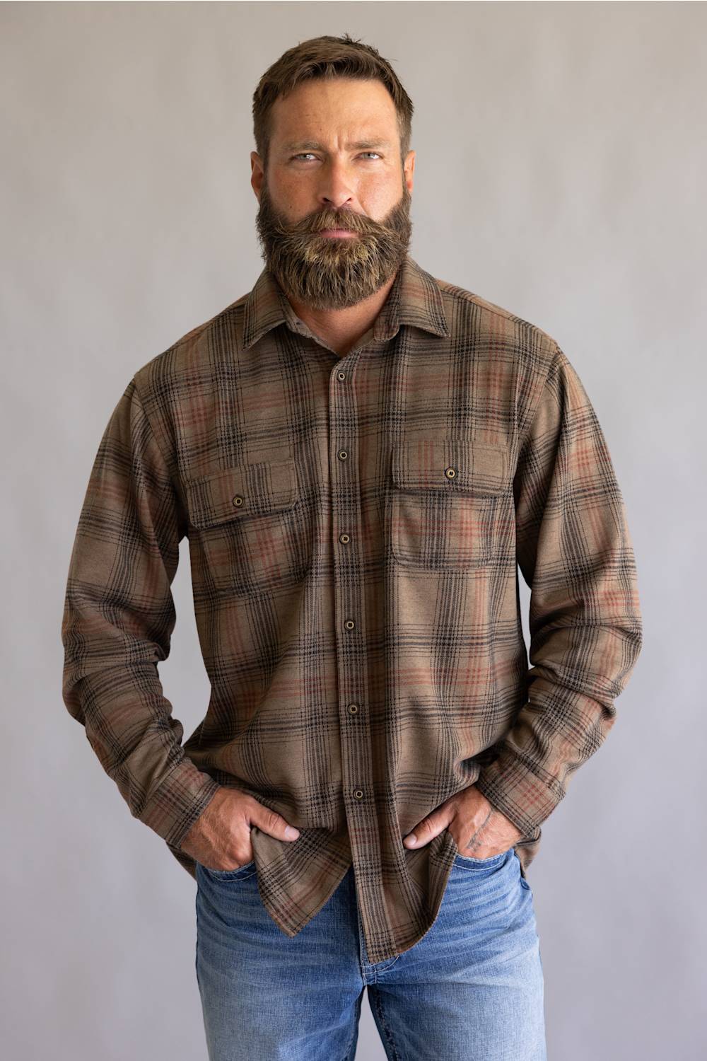 A man with a full beard wearing a brown and beige plaid button-down flannel shirt and blue jeans, standing against a neutral gray studio background.