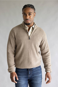 A man wearing a light beige ribbed quarter-zip pullover sweater over a cream collared shirt and blue jeans, standing against a neutral gray studio background.