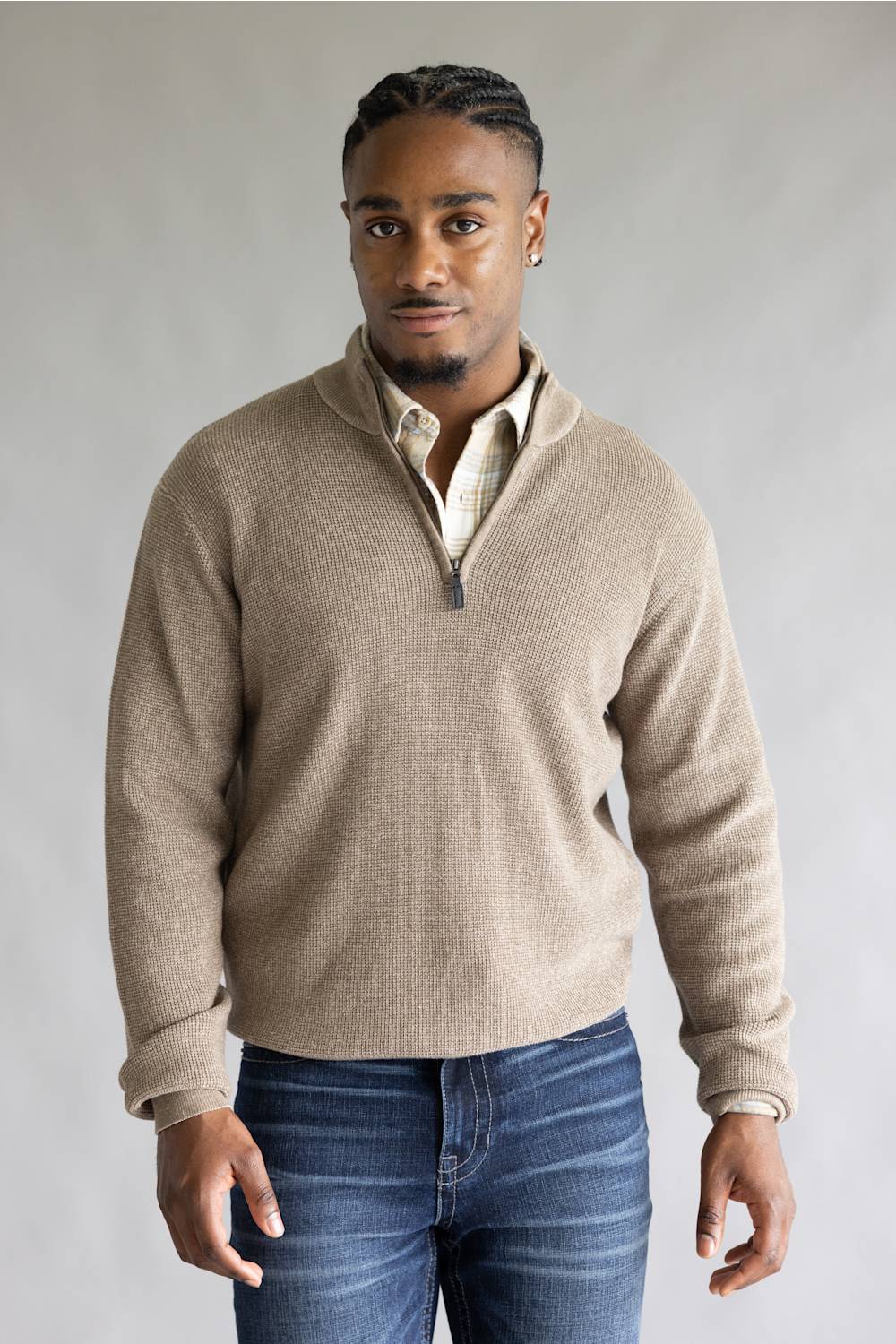 A man wearing a light beige ribbed quarter-zip pullover sweater over a cream collared shirt and blue jeans, standing against a neutral gray studio background.