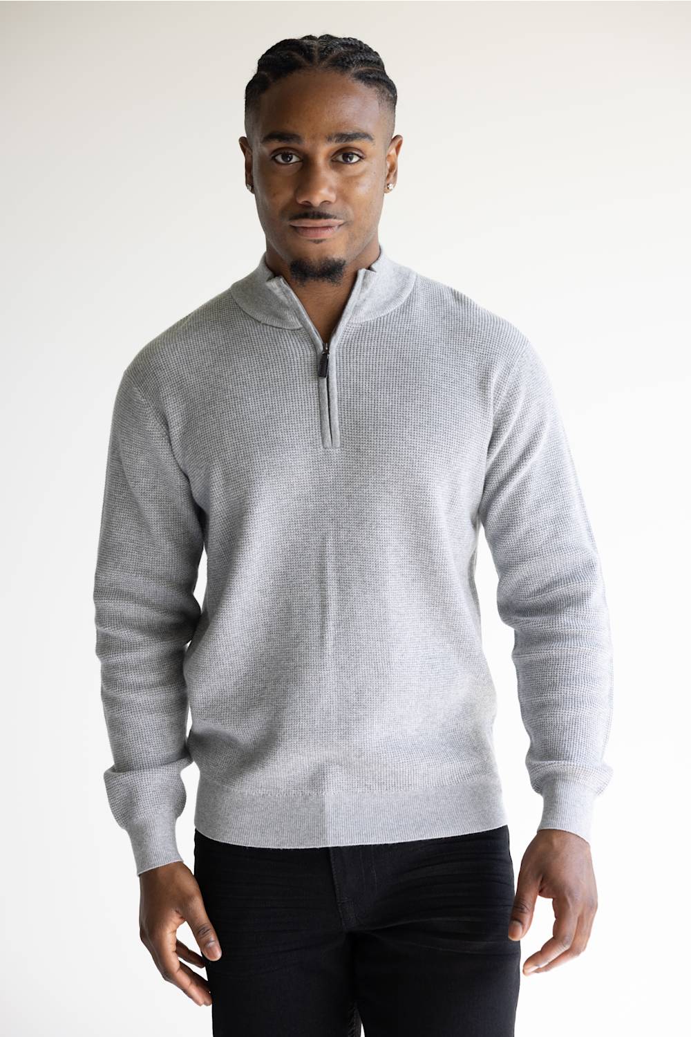 A man wearing a light gray quarter-zip knit sweater and black trousers, standing against a plain light gray studio background.