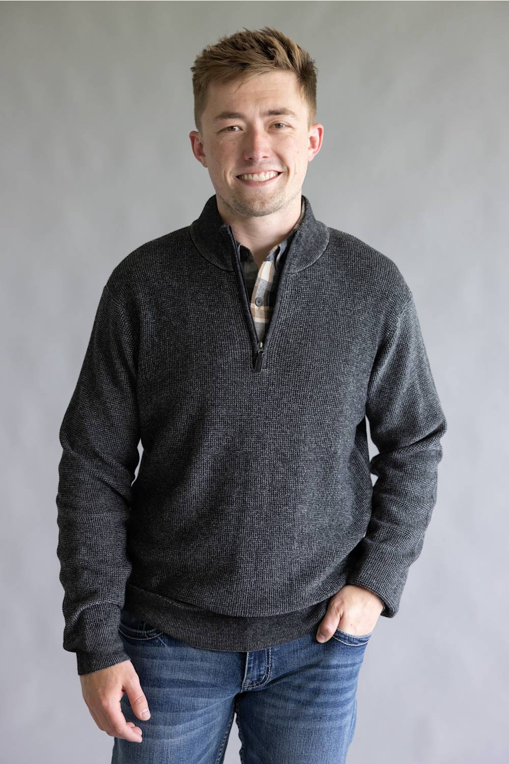A man wearing a dark gray quarter-zip knit sweater over a plaid shirt and blue jeans, standing against a neutral gray studio background.