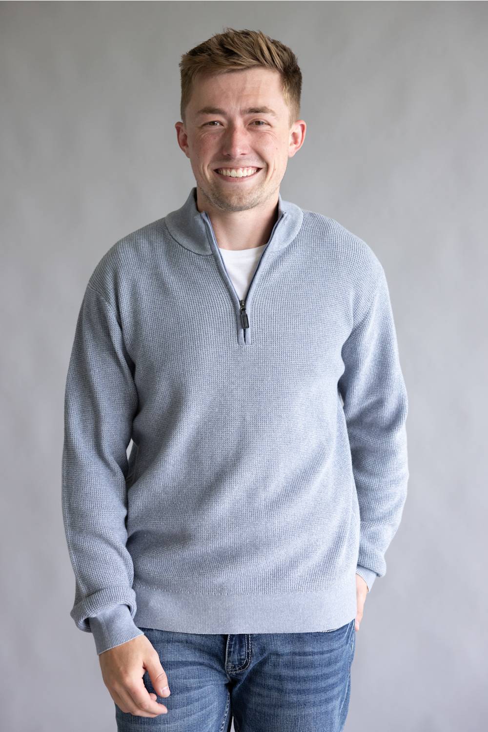 A smiling man wearing a light gray quarter-zip knit sweater over a white undershirt and blue jeans, standing against a neutral gray studio background.