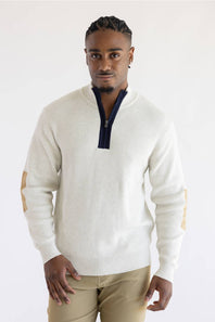 A man wearing a cream ribbed knit quarter-zip sweater with a dark navy collar and tan elbow patches, paired with khaki pants, against a plain white studio background.