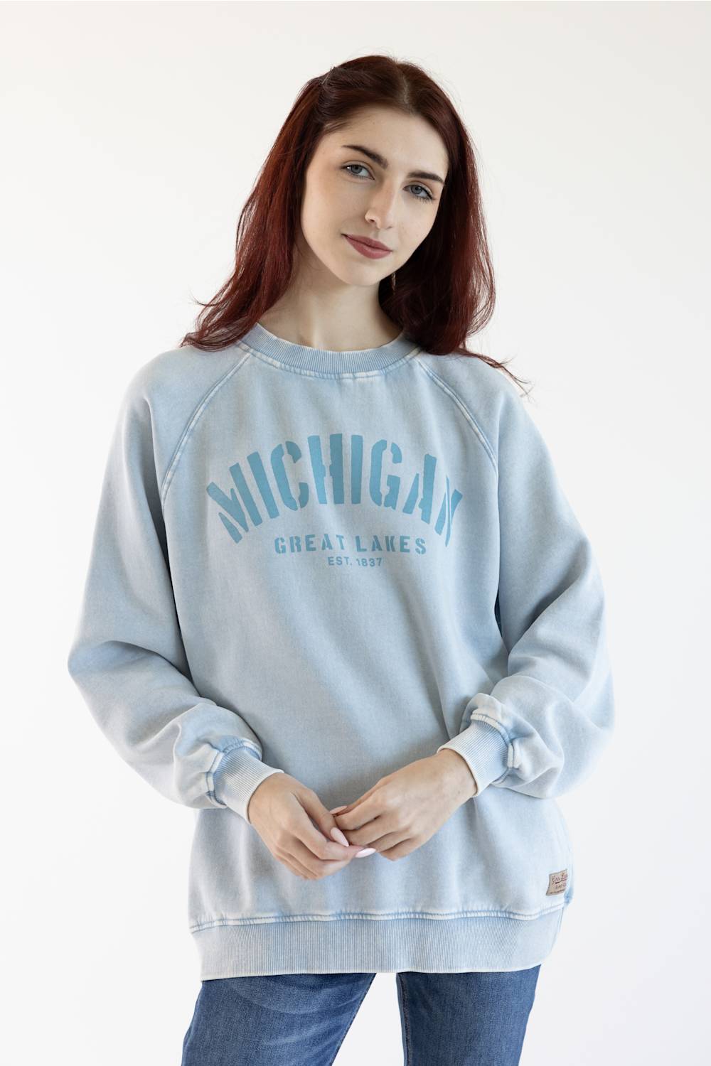 A woman with red hair wearing a light blue washed sweatshirt with 'MICHIGAN GREAT LAKES EST. 2017' printed in blue on the front, paired with blue jeans, against a plain white background.