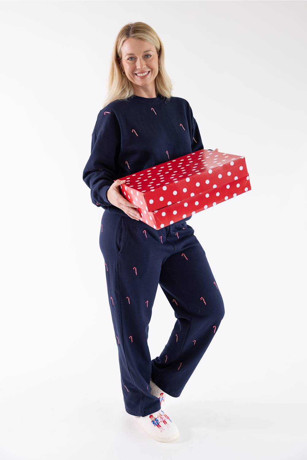 A woman smiling while holding a large red and white polka-dot gift box, wearing a navy blue crewneck fleece sweatshirt and matching sweatpants with an all-over pink and white candy cane embroidery pattern, and white sneakers.