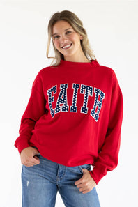 Front view of a smiling woman wearing the red 1897 Active Faith mock neck sweatshirt with navy blue "FAITH" applique lettering filled with white polka dots, paired with blue jeans against a plain white background.