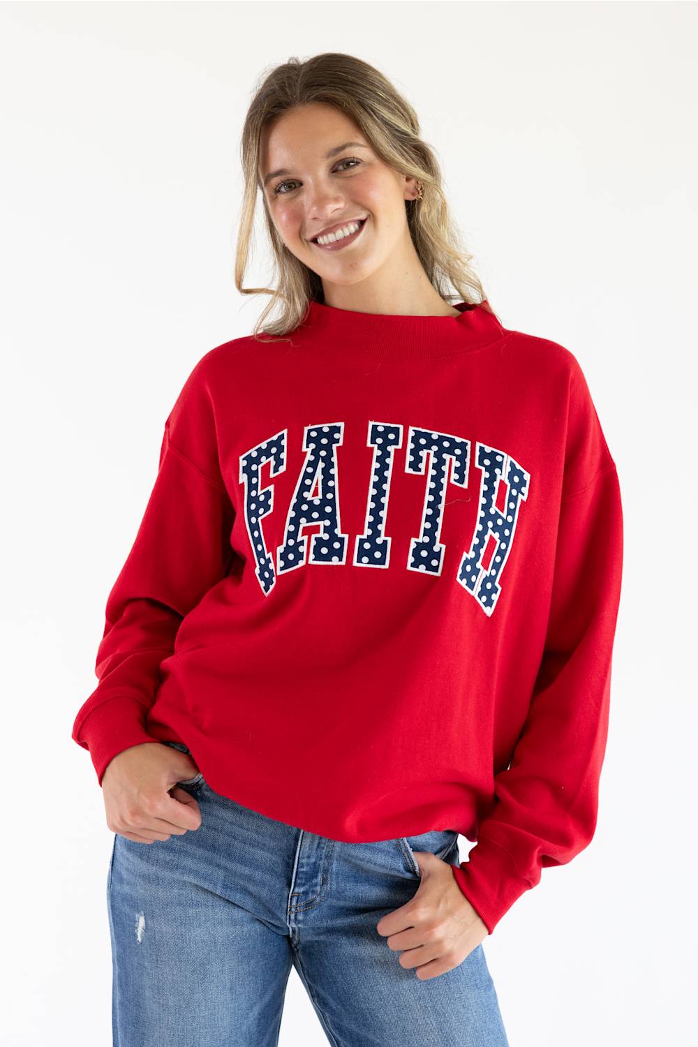 Front view of a smiling woman wearing the red 1897 Active Faith mock neck sweatshirt with navy blue "FAITH" applique lettering filled with white polka dots, paired with blue jeans against a plain white background.