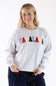 Studio portrait of a blonde woman wearing an 1897 Active Falalala Patch Embroidered Fleece Sweatshirt for Women in Ice Grey with colorful chenille lettering and dark blue holiday-patterned lounge pants.