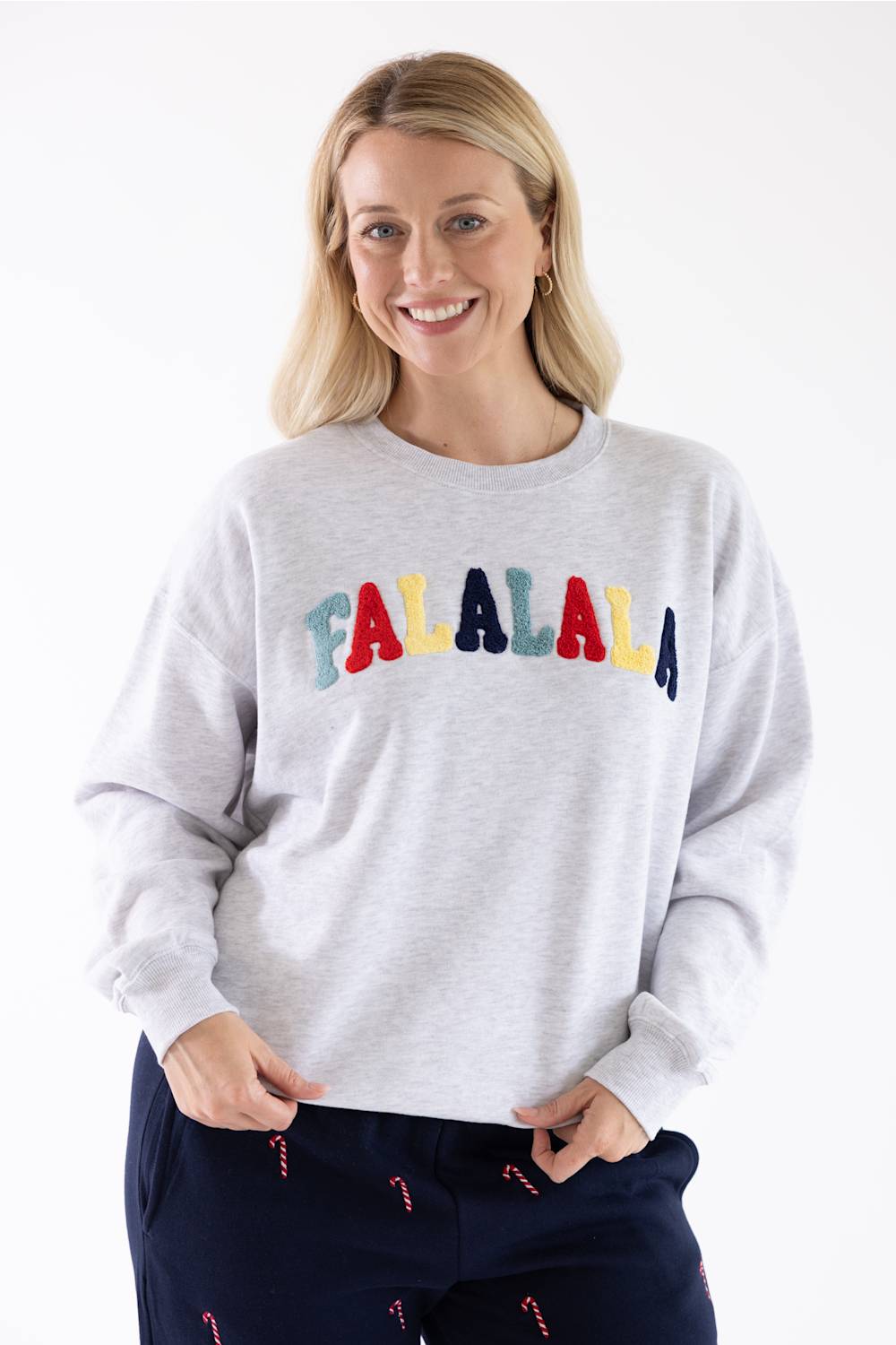 Studio portrait of a blonde woman wearing an 1897 Active Falalala Patch Embroidered Fleece Sweatshirt for Women in Ice Grey with colorful chenille lettering and dark blue holiday-patterned lounge pants.