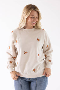 A smiling blond woman wearing a beige fleece crewneck sweatshirt with an all-over embroidered gingerbread man pattern.
