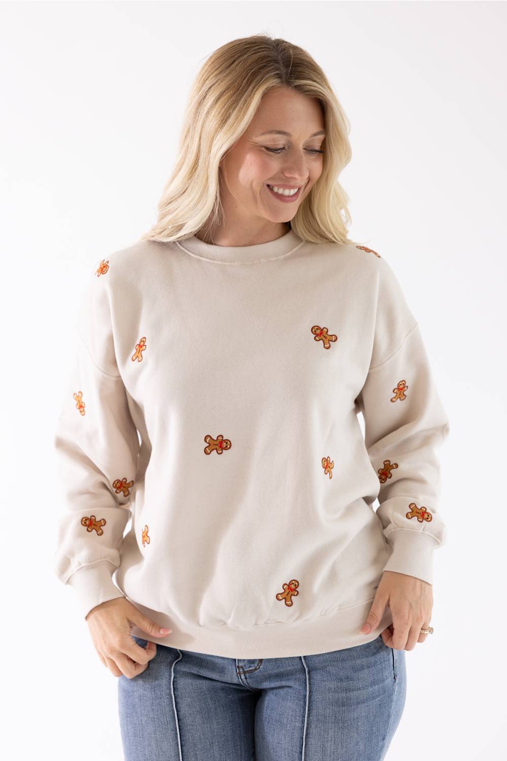 A smiling blond woman wearing a beige fleece crewneck sweatshirt with an all-over embroidered gingerbread man pattern.