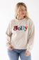 A smiling blond woman wearing a beige crewneck sweatshirt featuring multicolored chenille patch lettering that reads "Jolly" on the front.