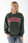 A smiling blond woman wearing a dark green crewneck fleece sweatshirt featuring red and white block lettering that reads "Merry" across the chest, and light wash jeans.