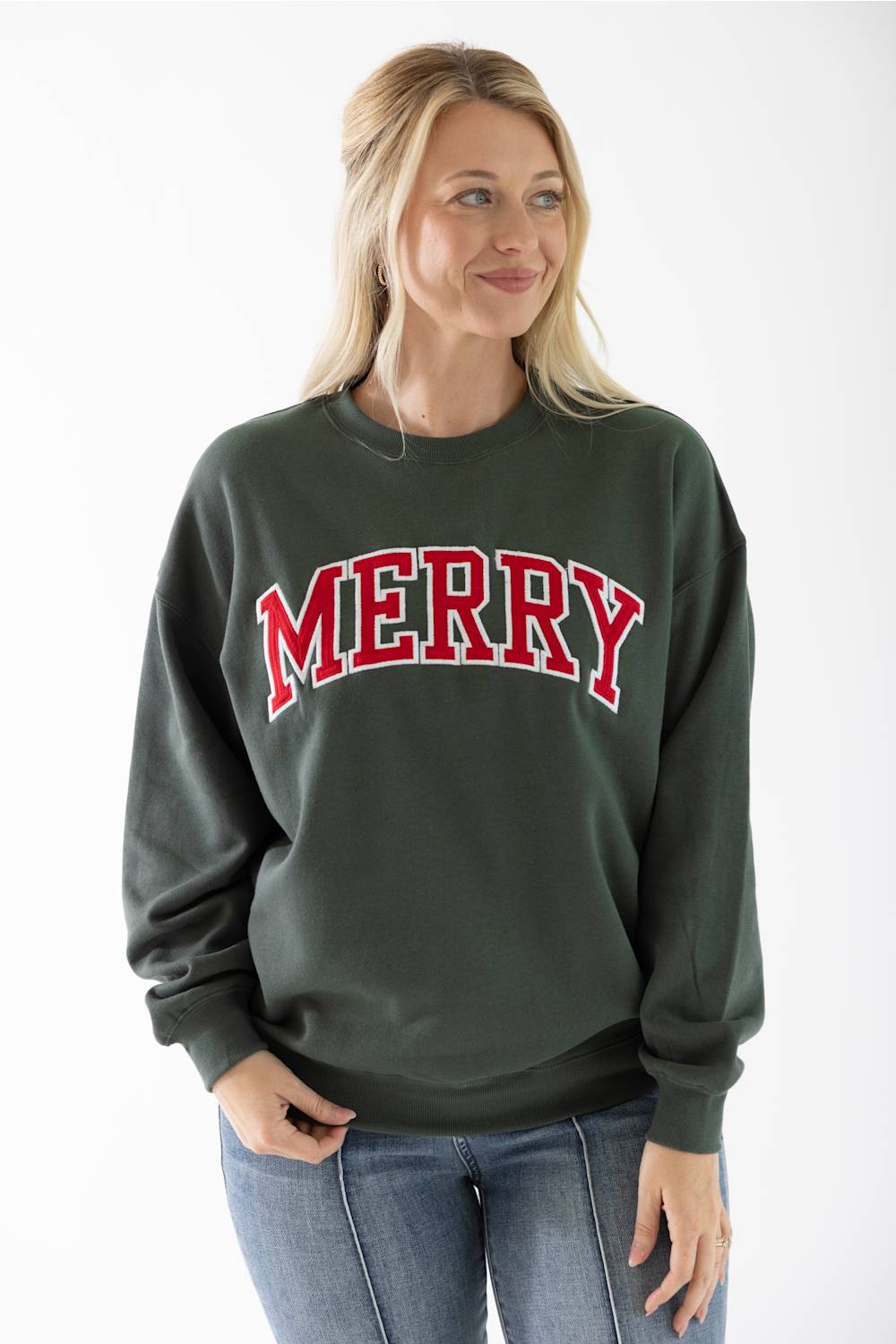 A smiling blond woman wearing a dark green crewneck fleece sweatshirt featuring red and white block lettering that reads "Merry" across the chest, and light wash jeans.