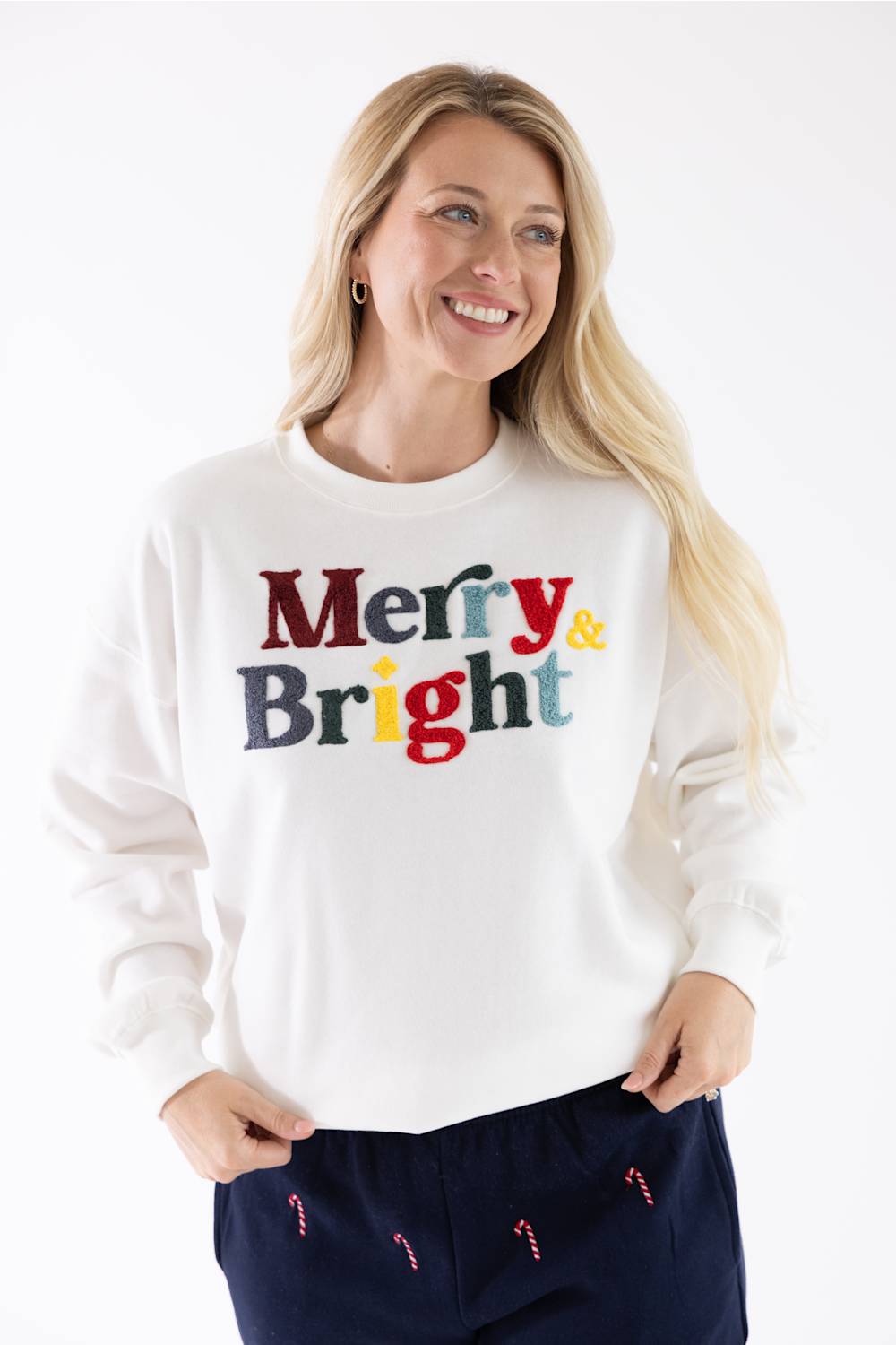 A smiling blonde woman wearing a white crewneck sweatshirt with colorful chenille "Merry & Bright" text across the chest, paired with dark blue lounge pants.