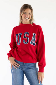 A woman wearing a red crewneck sweatshirt with 'USA' printed across the chest in white block letters filled with small white stars on a navy blue background, paired with blue jeans, against a plain white studio background.
