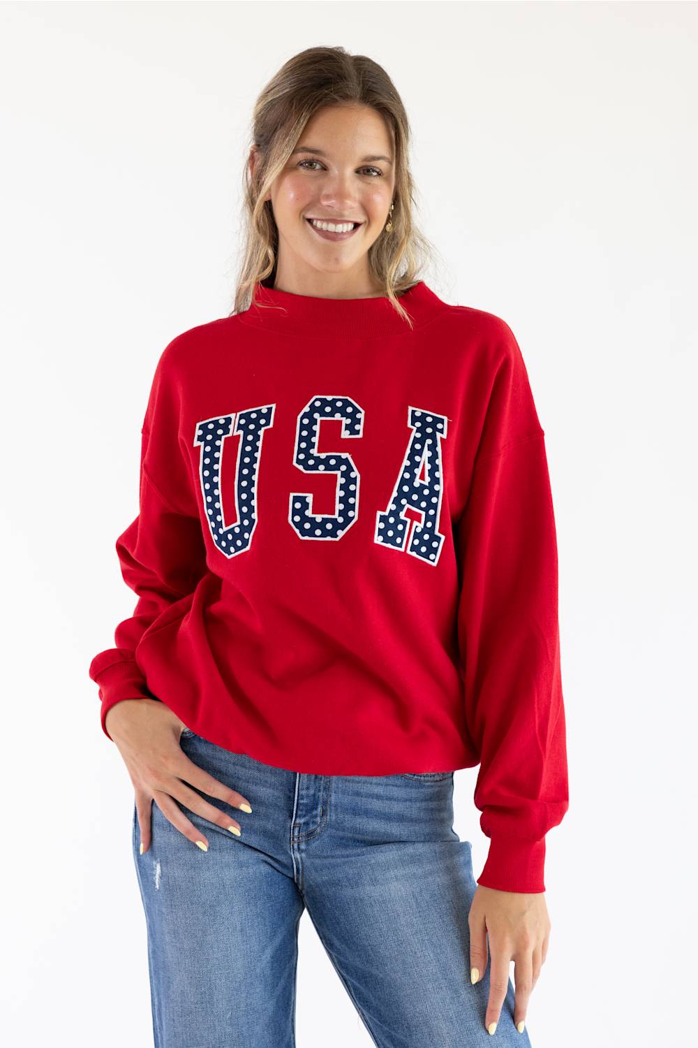 A woman wearing a red crewneck sweatshirt with 'USA' printed across the chest in white block letters filled with small white stars on a navy blue background, paired with blue jeans, against a plain white studio background.