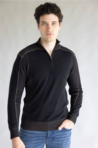 A man wearing a dark charcoal gray quarter-zip knit sweater with contrasting seam detailing on the shoulders and sleeves, paired with blue jeans, against a neutral gray background.