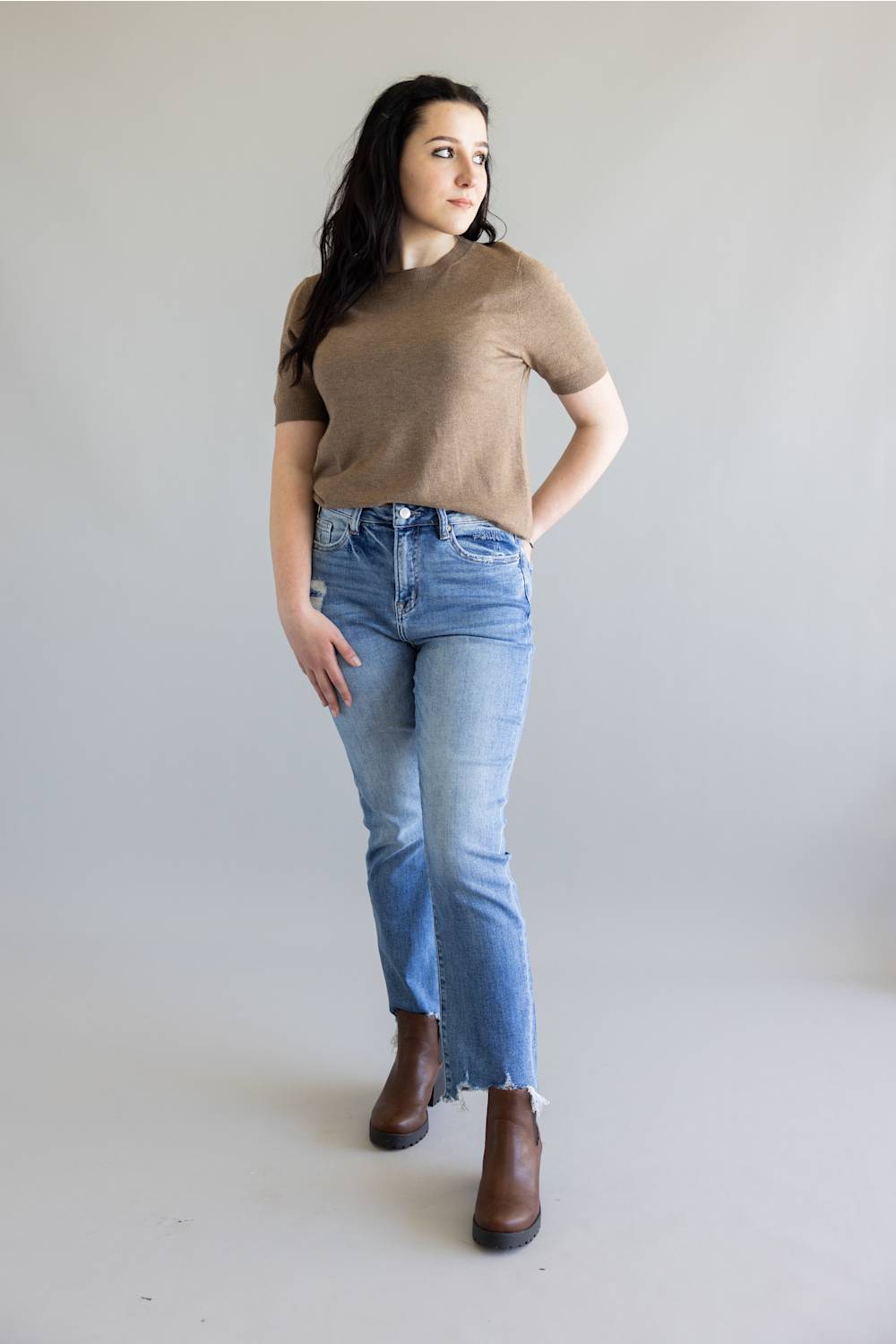 A woman modeling a brown short-sleeve knit top and light-wash blue jeans with brown ankle boots, standing against a plain light gray studio background.