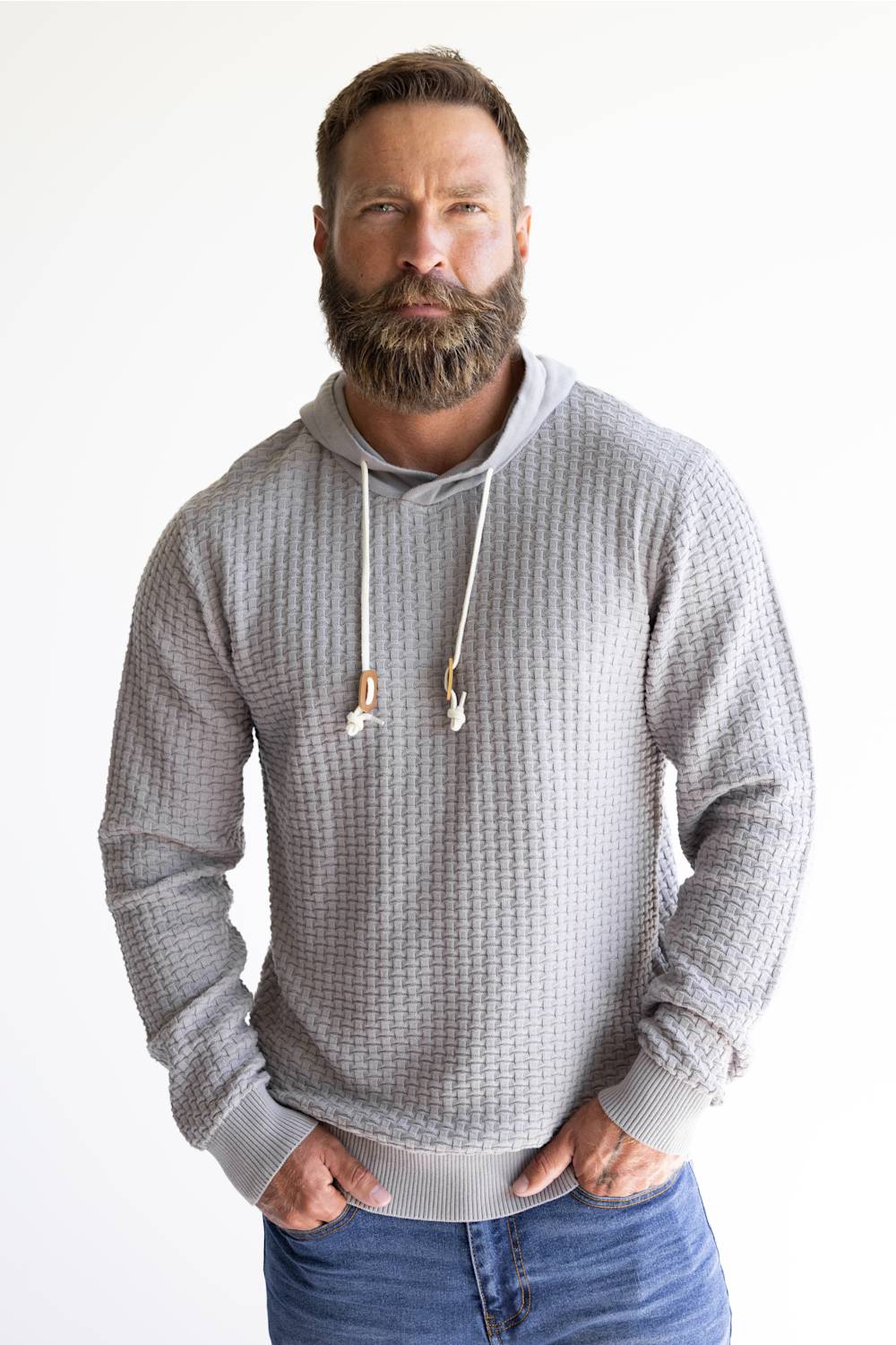 A man with a full beard wearing a light gray waffle-knit hoodie with white drawstrings and blue jeans, standing against a plain white background.