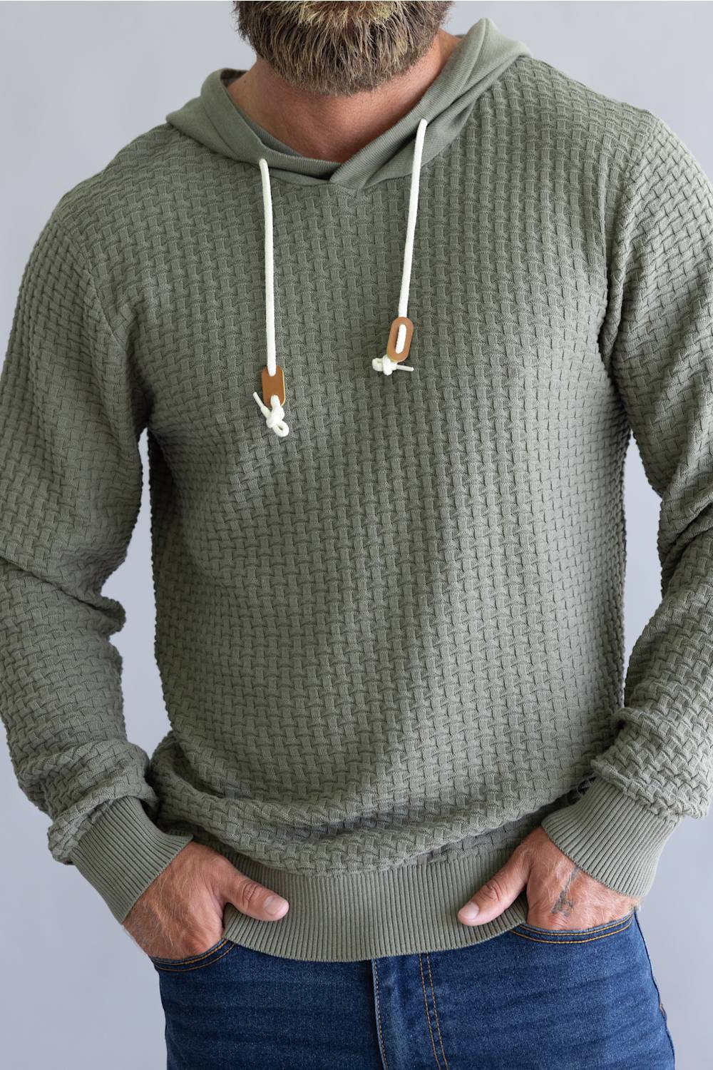 A man wearing an olive green waffle-knit hoodie with white drawstrings, paired with blue jeans, shown from the waist up against a neutral gray background.