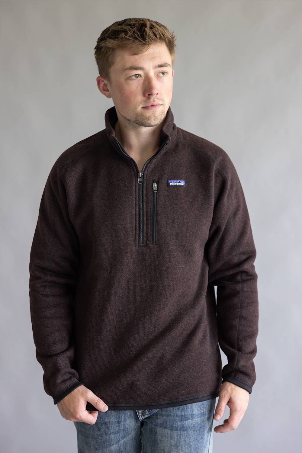 A man wearing a dark brown Patagonia quarter-zip fleece pullover sweater and blue jeans, standing against a neutral gray studio background.