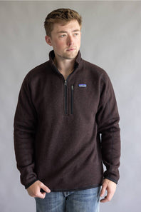 A man wearing a dark brown Patagonia quarter-zip fleece pullover sweater and blue jeans, standing against a neutral gray studio background.
