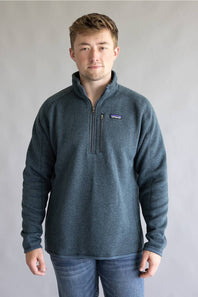 A man wearing a heathered slate blue quarter-zip fleece pullover with the Patagonia logo on the chest, paired with blue jeans, standing against a neutral gray studio background.