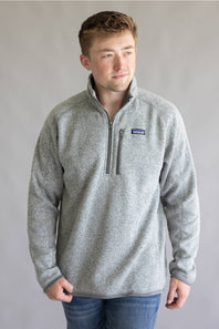 A man wearing a heather gray quarter-zip fleece pullover with a small Patagonia logo on the left chest, paired with blue jeans, standing against a neutral gray studio background.