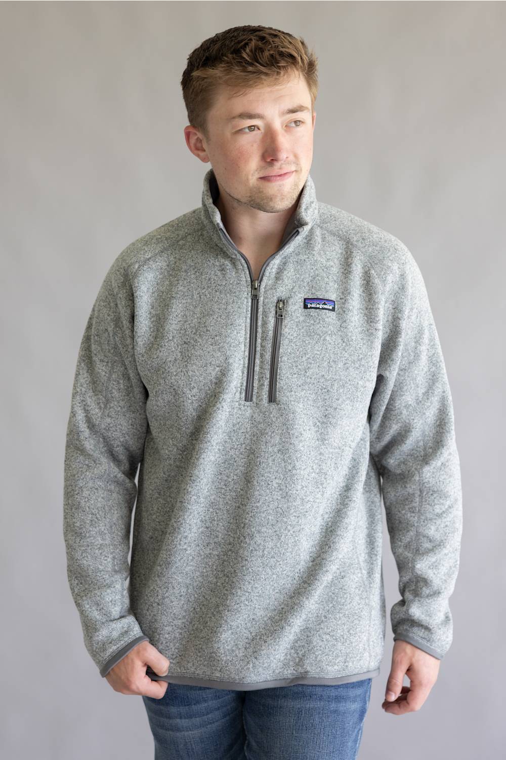 A man wearing a heather gray quarter-zip fleece pullover with a small Patagonia logo on the left chest, paired with blue jeans, standing against a neutral gray studio background.