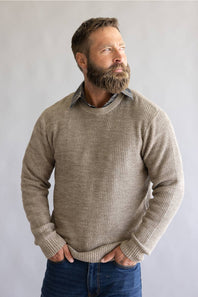 A man with a full beard wearing a textured, waffle-knit crewneck sweater in a taupe-beige color, layered over a collared shirt and paired with blue jeans, standing against a neutral gray background.