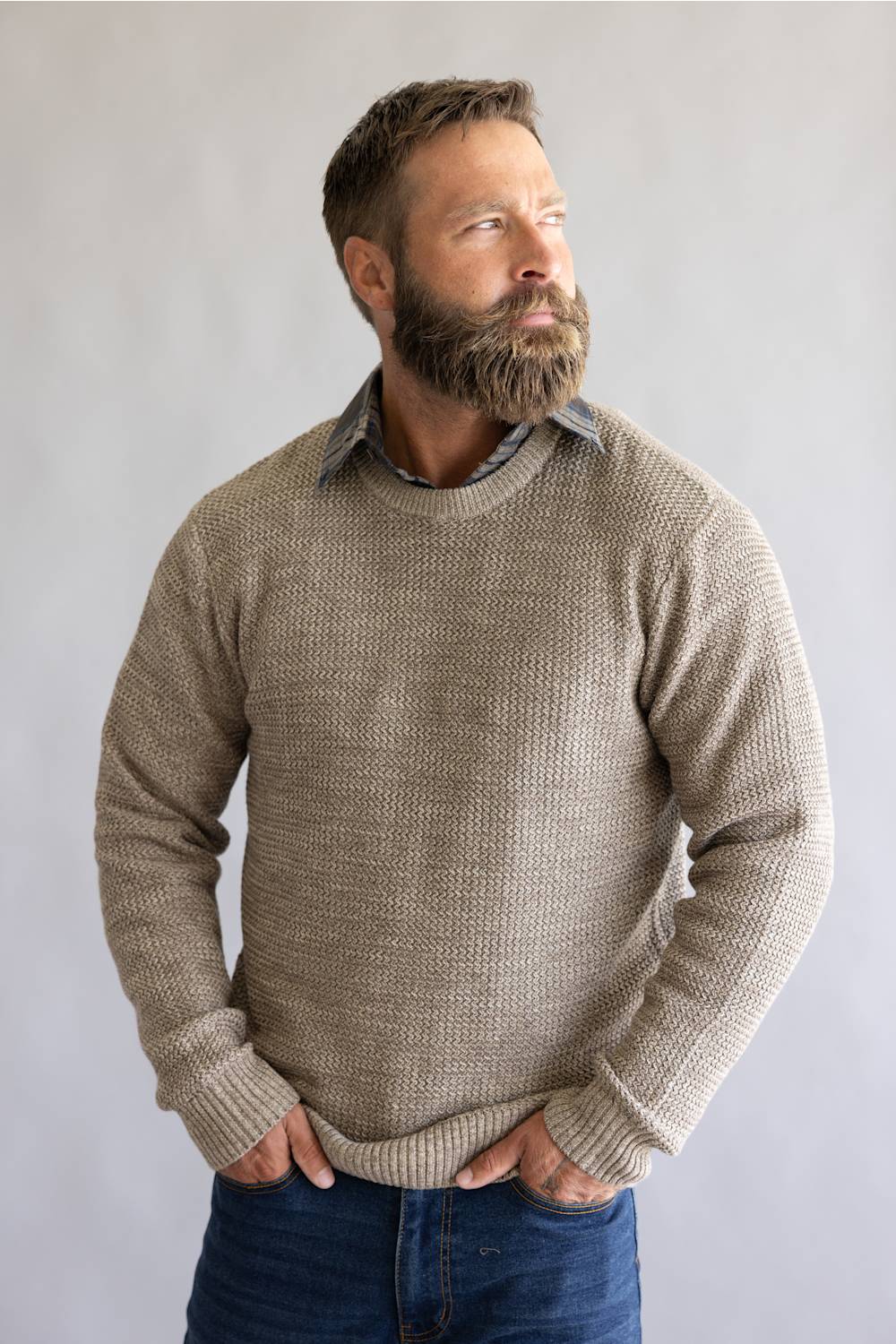 A man with a full beard wearing a textured, waffle-knit crewneck sweater in a taupe-beige color, layered over a collared shirt and paired with blue jeans, standing against a neutral gray background.