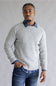 A man wearing a light gray textured knit sweater over a blue and white checkered collared shirt, paired with blue jeans, standing against a neutral gray background.