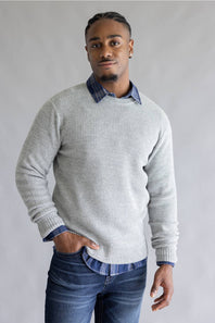 A man wearing a light gray textured knit sweater over a blue and white checkered collared shirt, paired with blue jeans, standing against a neutral gray background.
