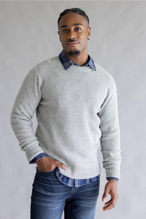 A man wearing a light gray textured knit sweater over a blue and white checkered collared shirt, paired with blue jeans, standing against a neutral gray background.