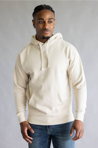 A man wearing a cream-colored pullover hoodie with drawstrings and a front kangaroo pocket, paired with blue jeans, standing against a neutral gray studio background.