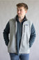 A man wearing a gray and navy fleece vest over a matching quarter-zip pullover sweater and blue jeans, standing against a plain light gray background. The vest features a small Patagonia logo patch on the left chest.